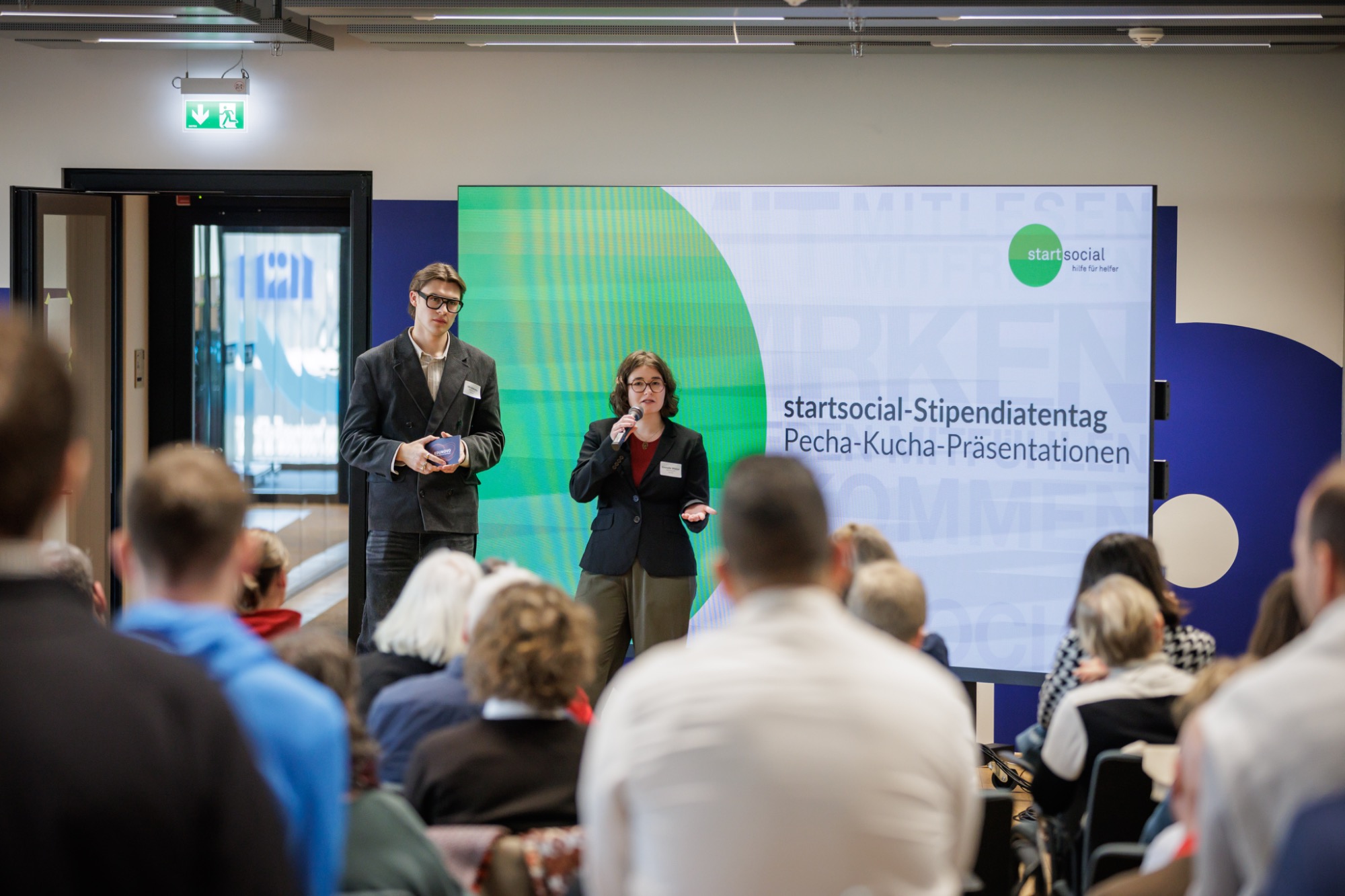 Vorstellung von Edunovo auf startsocial-Stipendientag Zwei Personen präsentieren auf einer Bühne vor Publikum, im Hintergrund ein großes Display.